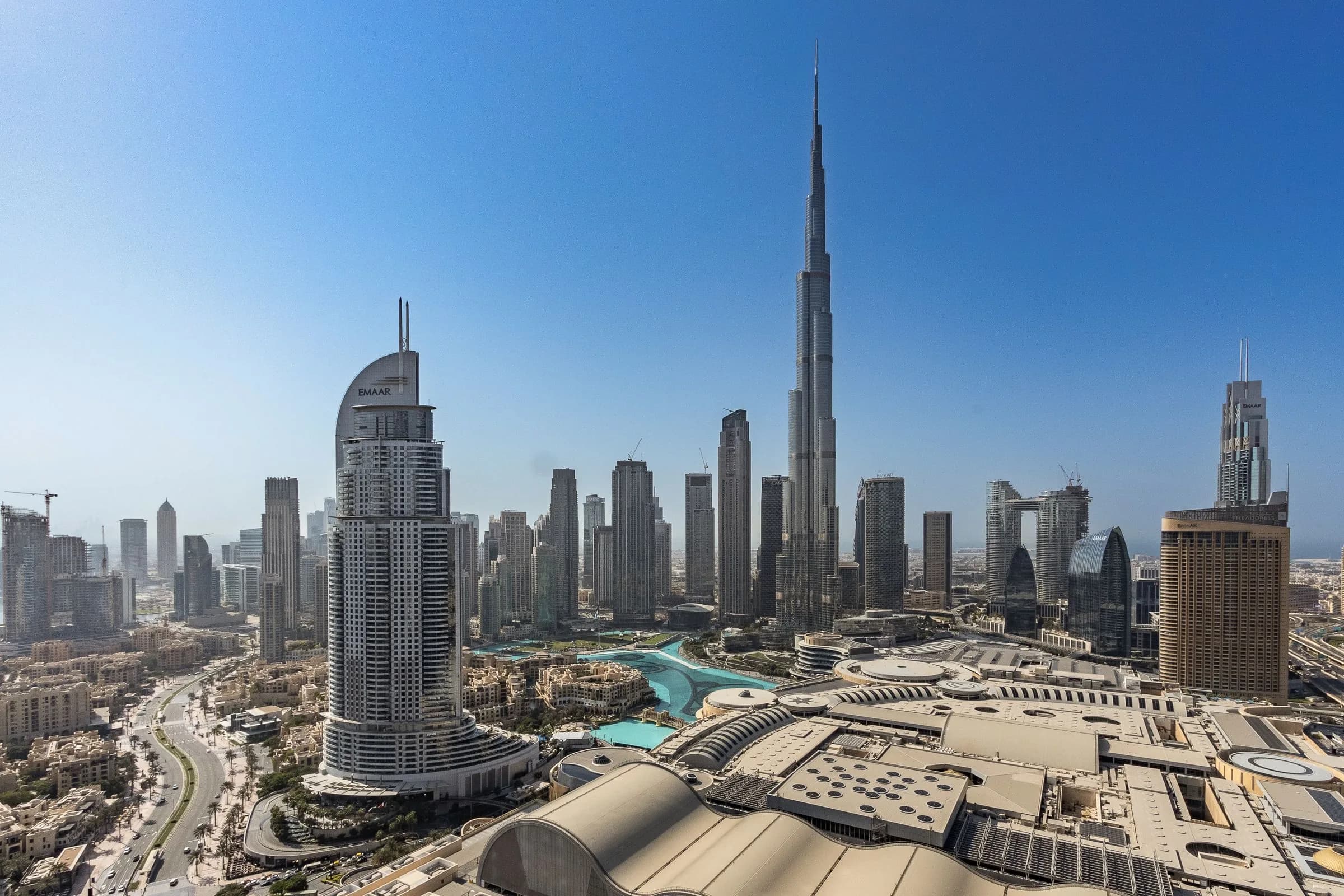 An aerial view of Downtown Dubai with the Burj Khalifa rising above the skyline and the roofs of Dubai Mall, under a clear blue sky.